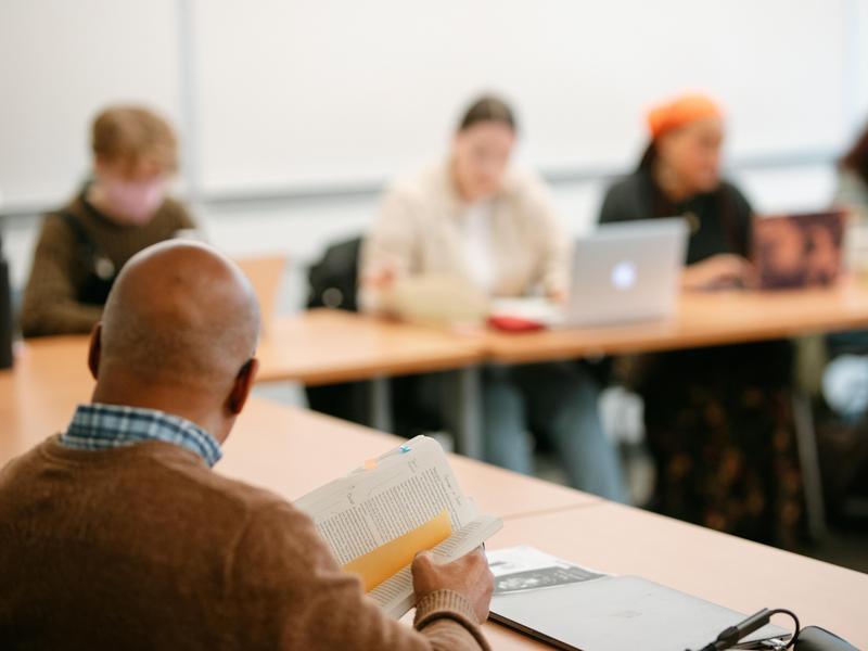 Over-the-shoulder view of a professor holding an open book while teaching students in a classroom.