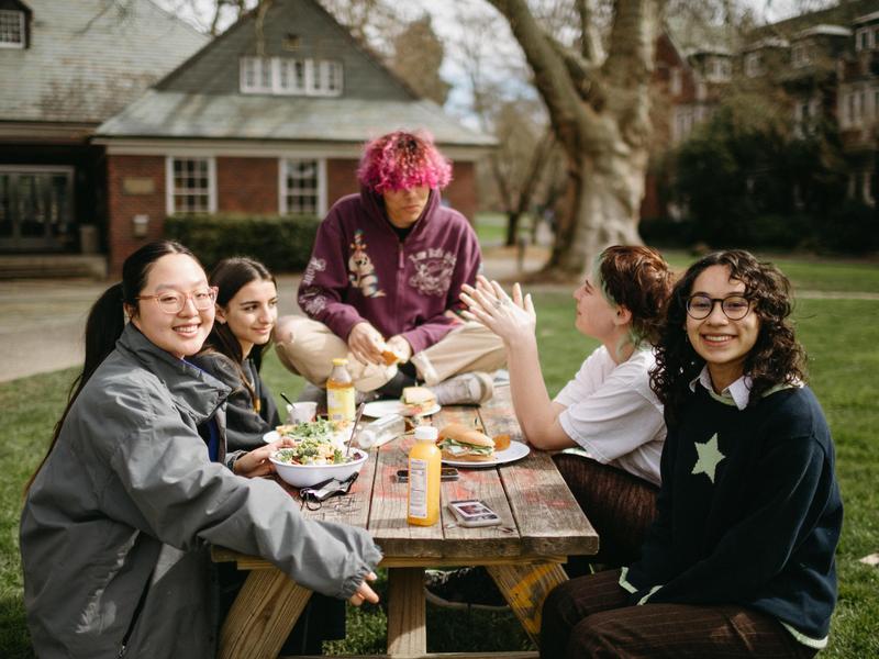 Five students eating lunch outside, with two smiling at the camera while the others talk.