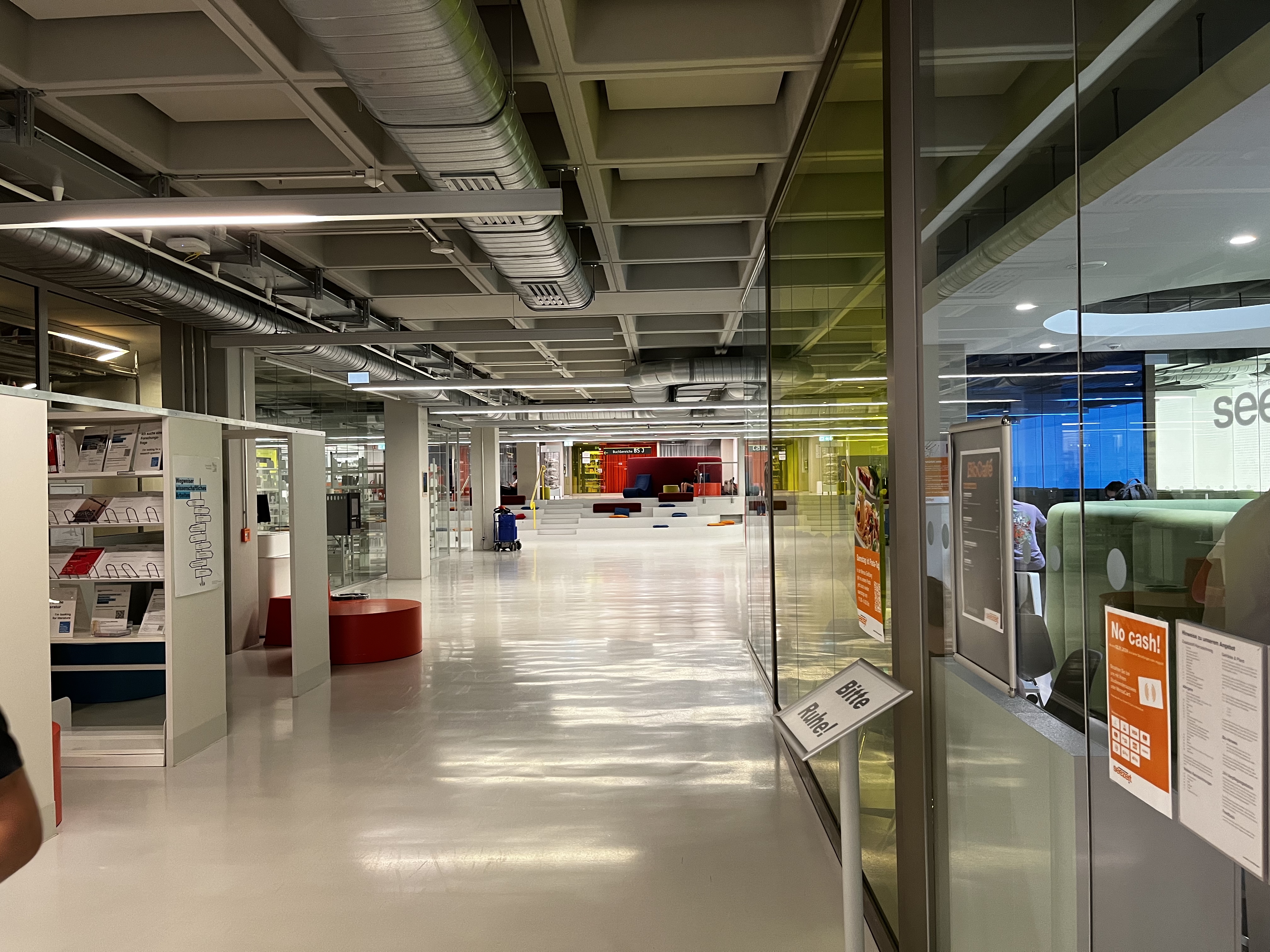 Hallway within the university. Bright, with white floors and bookshelves, and a glass wall to the right.
