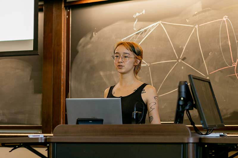 A student gives a talk in front of a projection screen in a wood paneled room.