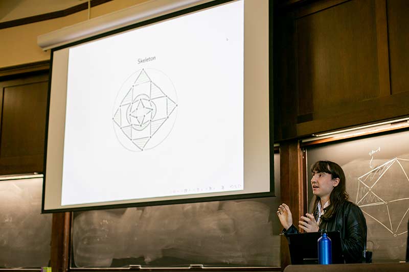 A student gives a talk in front of a projection screen in a wood paneled room.