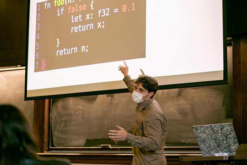 A student gives a talk in front of a projection screen in a wood paneled room.