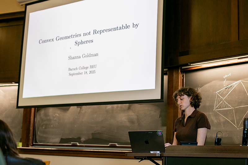 A student gives a talk in front of a projection screen in a wood paneled room.