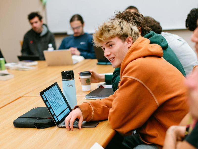 Students in a classroom sitting around a rectangular table with laptops and books.