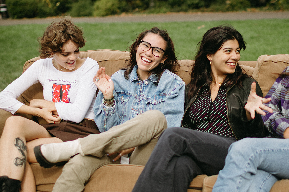 Three students laughing while chatting and sitting on a couch placed on the Great Lawn.