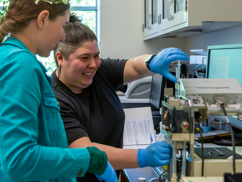 A professor and student smile while operating equipment in a science lab.