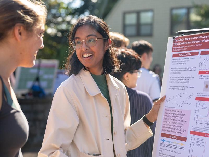 A student in a lab coat presents a research poster outside.