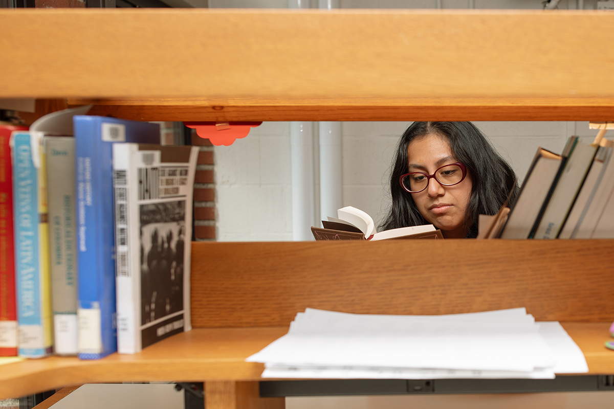 Flor Lopez reading a book behind a bookshelf.