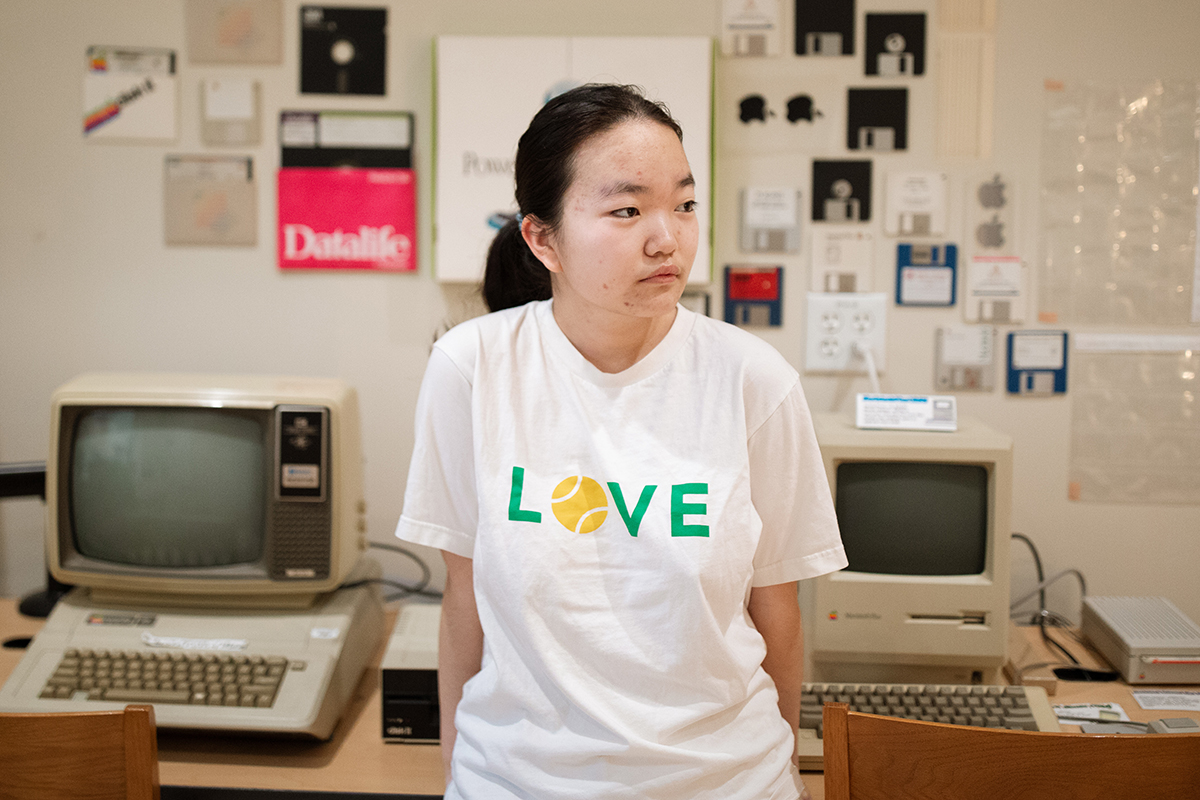 Luna Lu leaning against a desk that has two old computers.