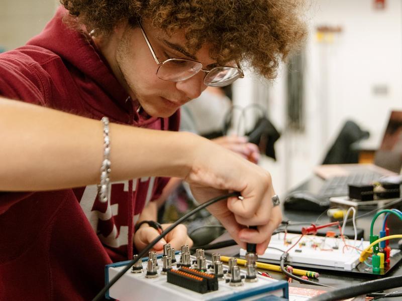 A student plugs a wire into an electronic device in a physics lab.