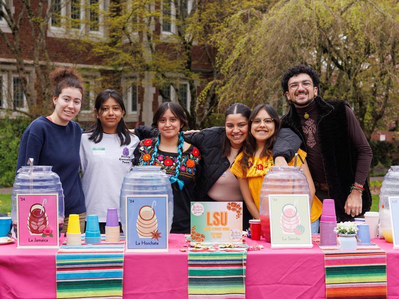 Six students pose behind a beverage table at an outdoor multicultural festival.
