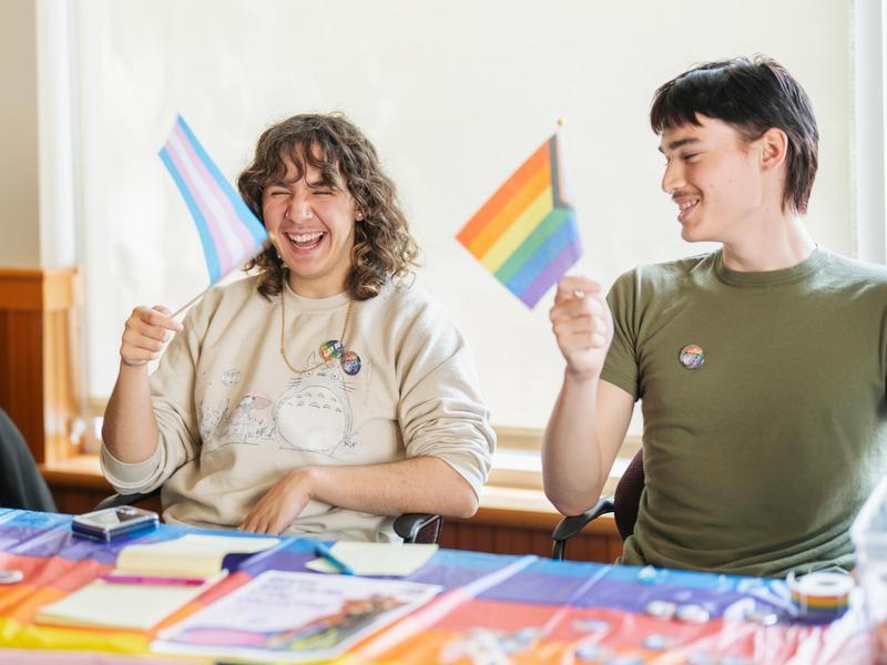 Two students smile while waving pride flags.