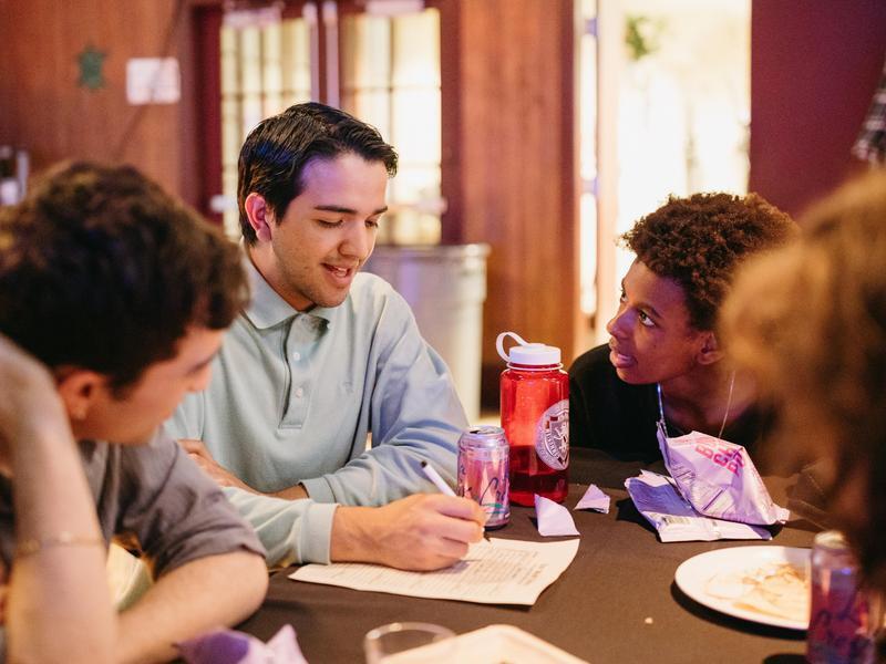 Three people sitting at a round table working together while one person writes on a piece of paper.