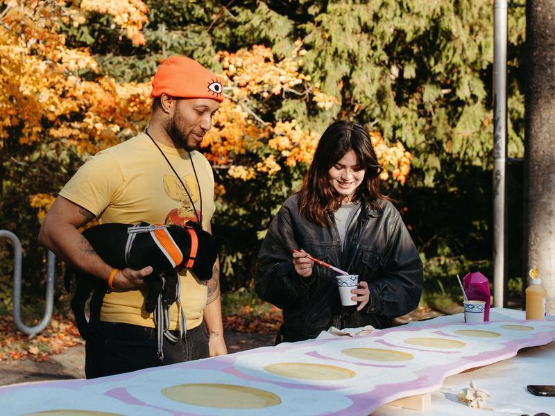 Two people painting a large plywood artwork outside.