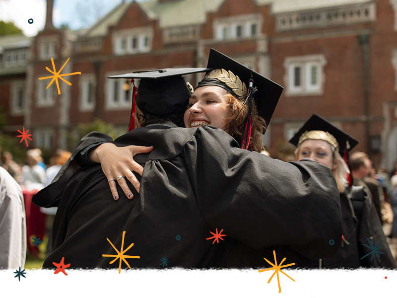 Two graduates in regalia embrace outside of Eliot Hall after Commencement.