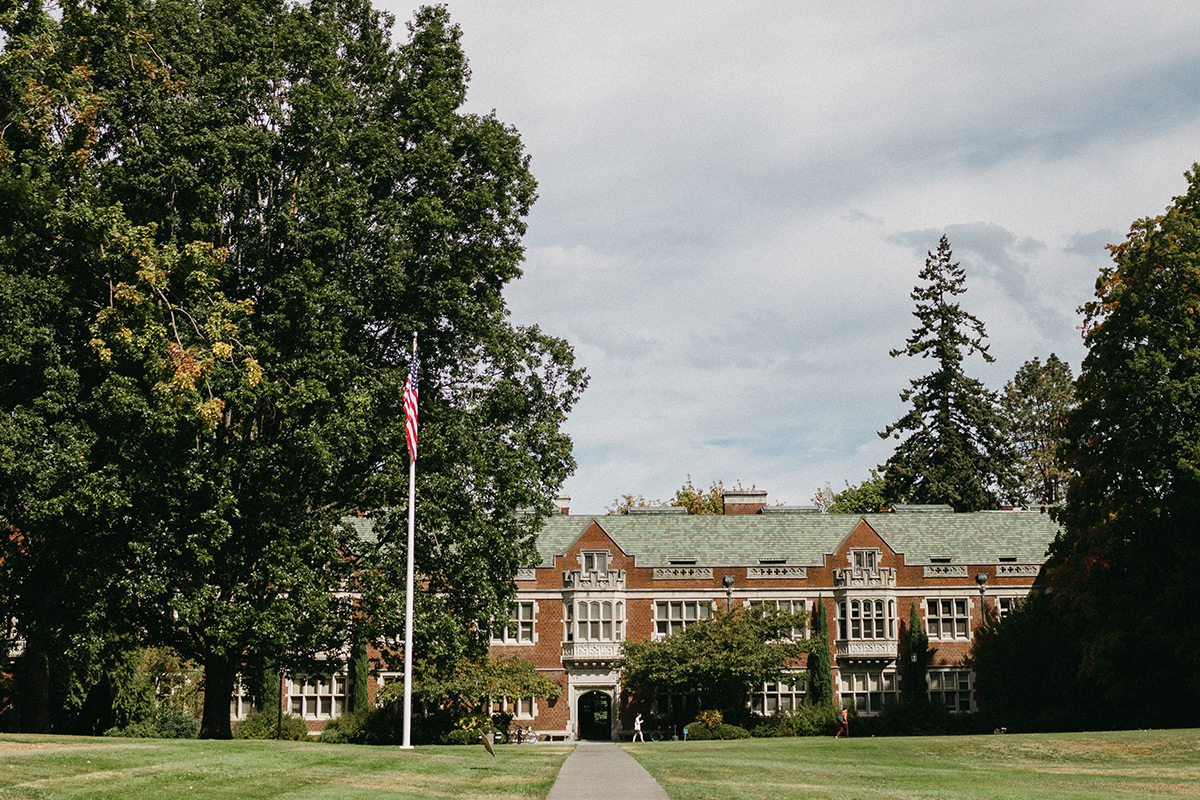 The front lawn on a sunny summer day leading up to Eliot Hall with a flag pole in the foreground carrying the American flag.