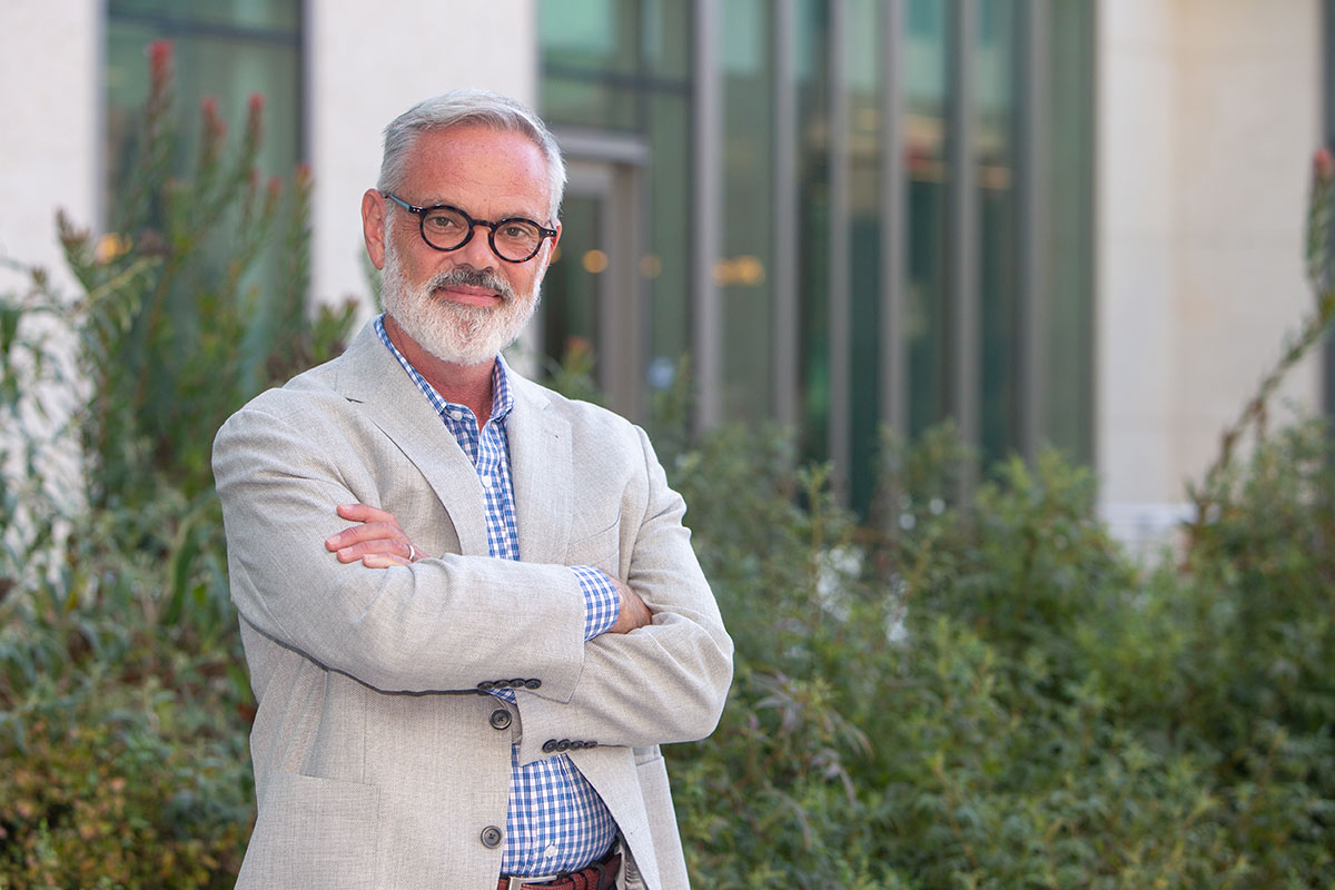 Headshot of man who wears black rimmed glasses, a beard, and a blazer