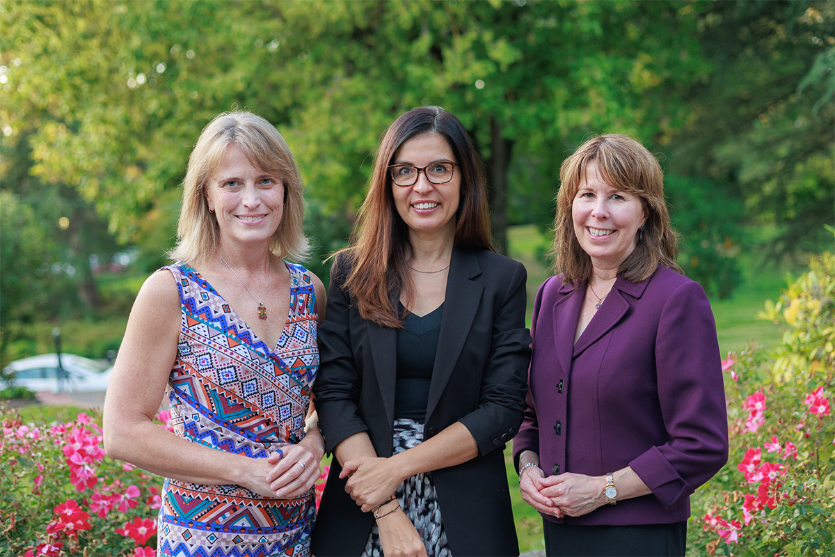 Three women pose, smiling, in front of greenery on campus. 