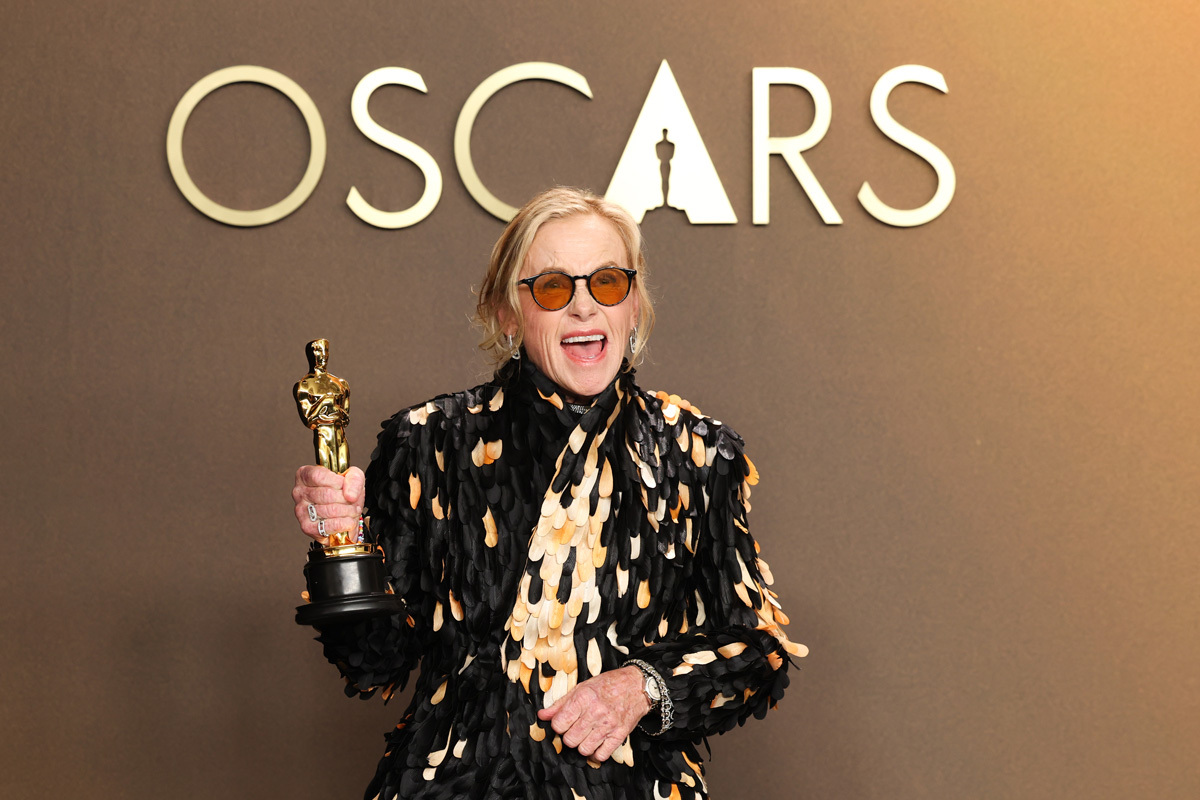 Amy Madigan stands in front of the Oscars logo holding her Oscar and smiling.