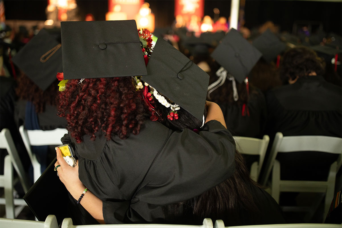 Two students wearing graduation caps embrace at commencement. 