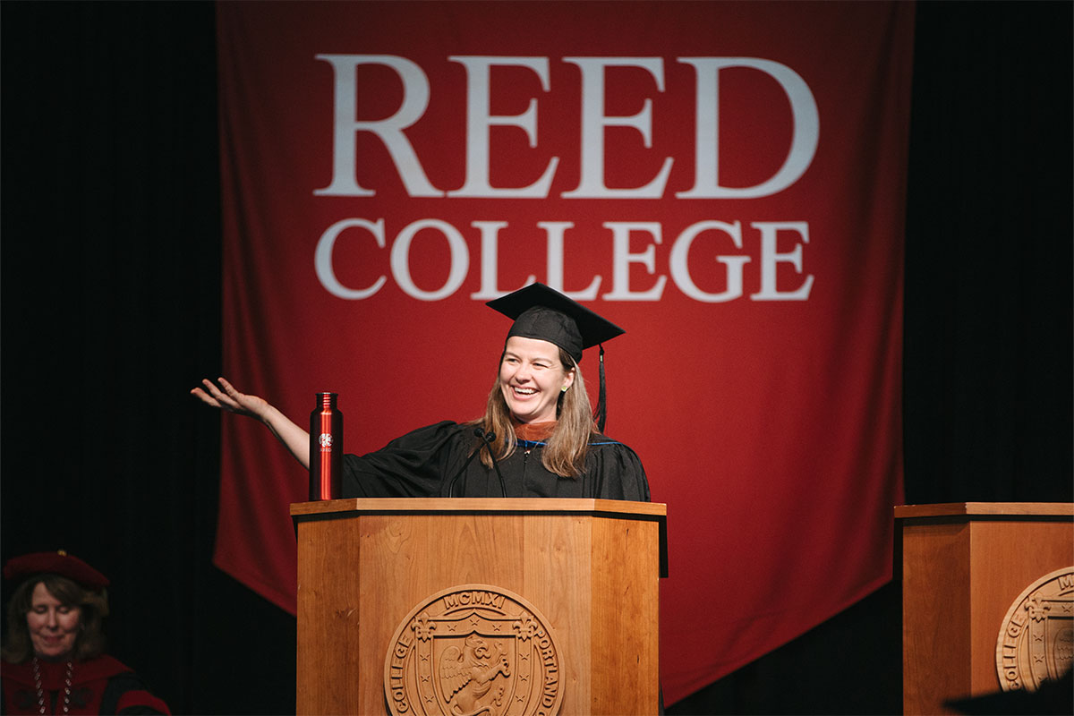 A woman speaks at a podium in front of a banner reading "Reed College."