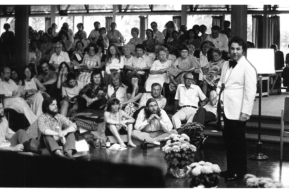 1980s era audience seated around violinist in a white coat and black bowtie.