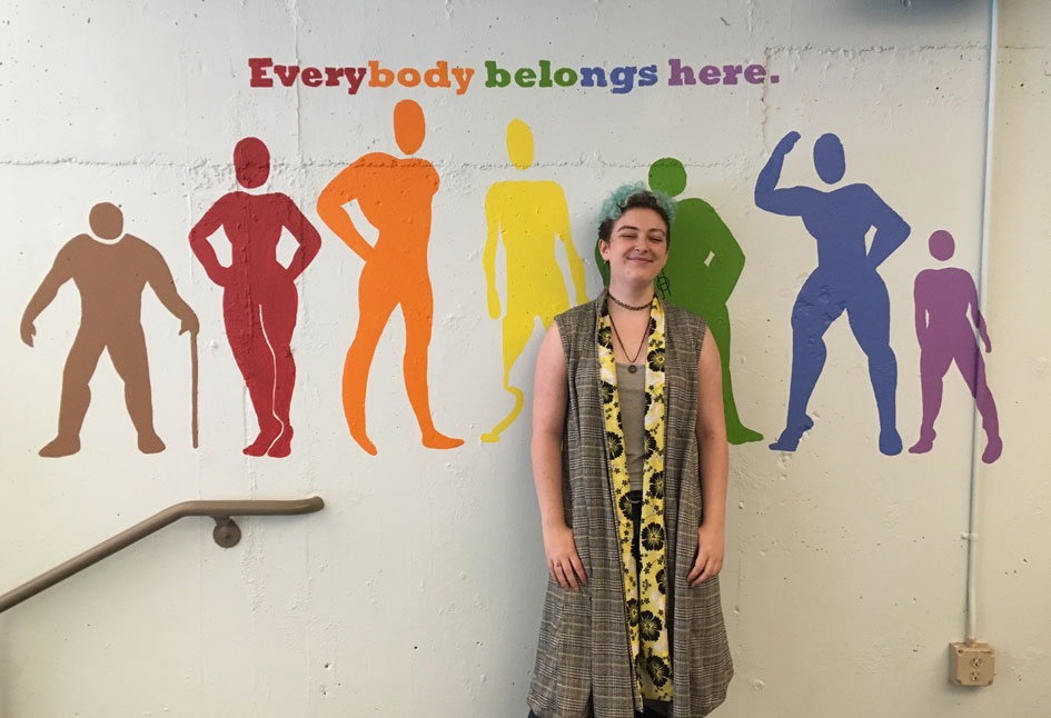 Lune Whitlock &amp;#8217;20 stands in front of their mural, &amp;#8220;Everybody Belongs Here,&amp;#8221; in the sports center weight room.
