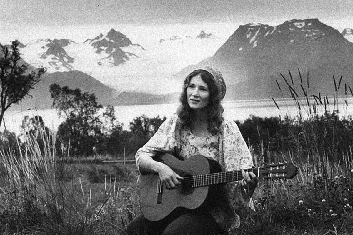 black and white photo of Mossy holding a guitar in front of mountains and a lake