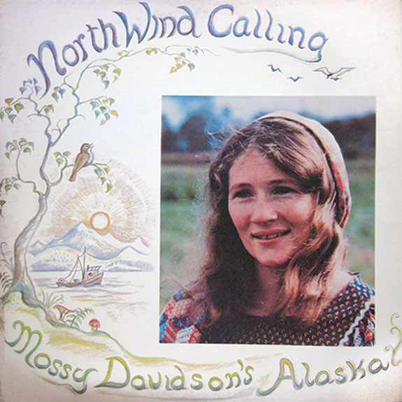 Lp cover with a photo of mossy. The cover says north wind calling at the top and mossy Davison’s Alaska at the bottom. It depicts a tree with mountains and a lake with about in the background.
