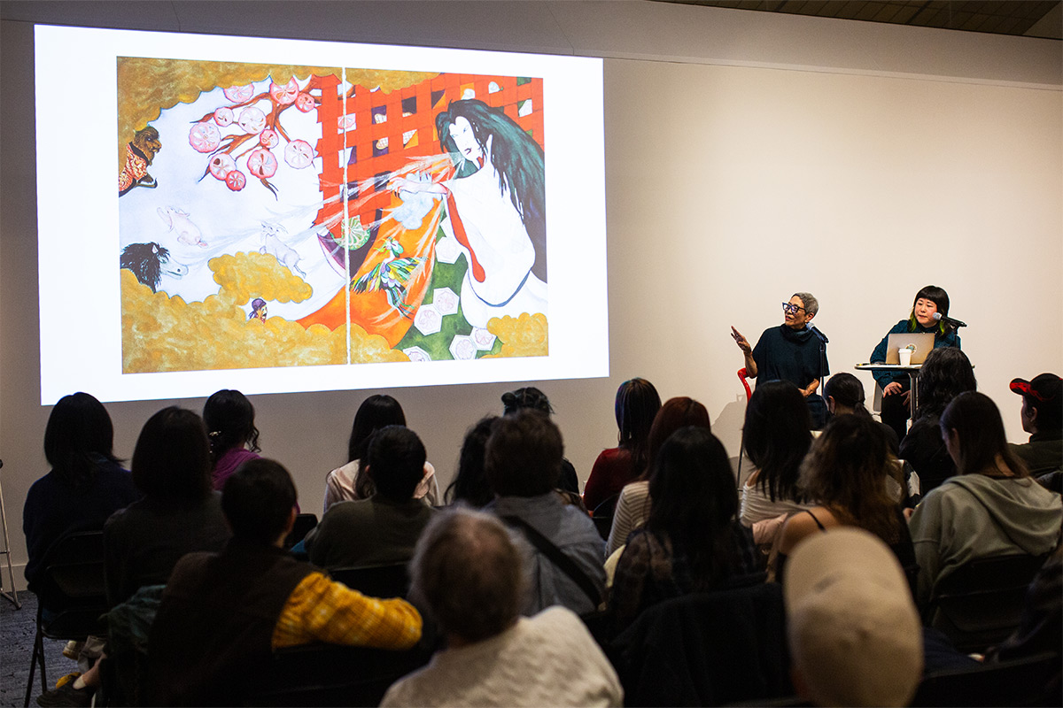 An audience watches as a woman gives a talk about her art displayed on the screen.