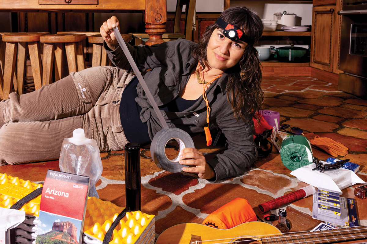 Athena sitting with ukulele on kitchen floor surrounded by items like maps, passport, tape, bags, sleeping mat