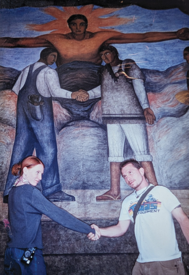 Rebecca and Noah holding hands in front of a wall painting of two figures holding hand