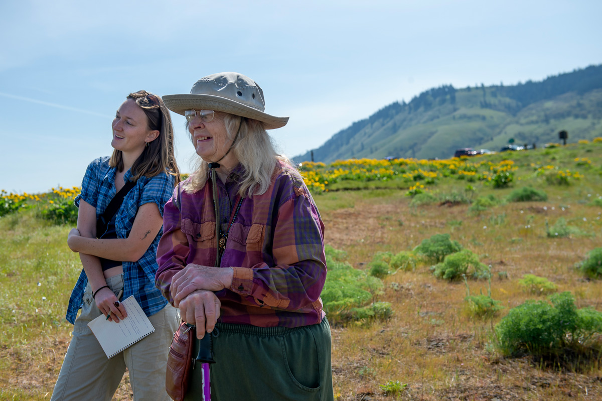 Two women stand smiling together, looking out at the landscape of the wildflowers surrounding them.