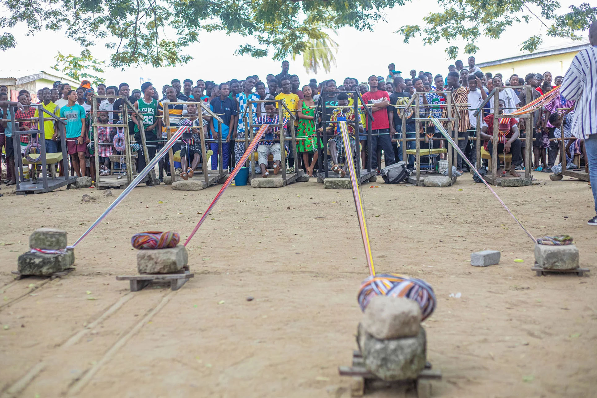 A crowd gathers to watch an outdorr weaving competition with fibers anchored by large rocks and spanning to looms a dozen feet away