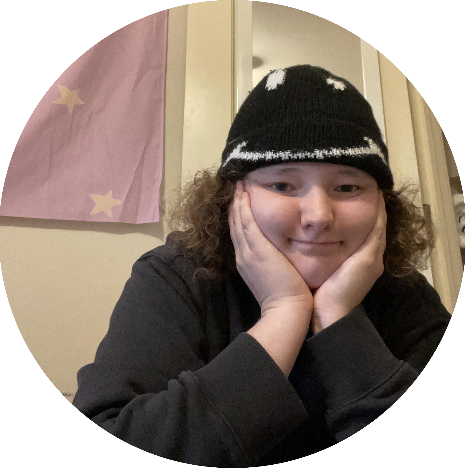 A light-skinned person with curly hair, in a black knitted cap with a monster design, rests their face in their hands and looks at the camera.