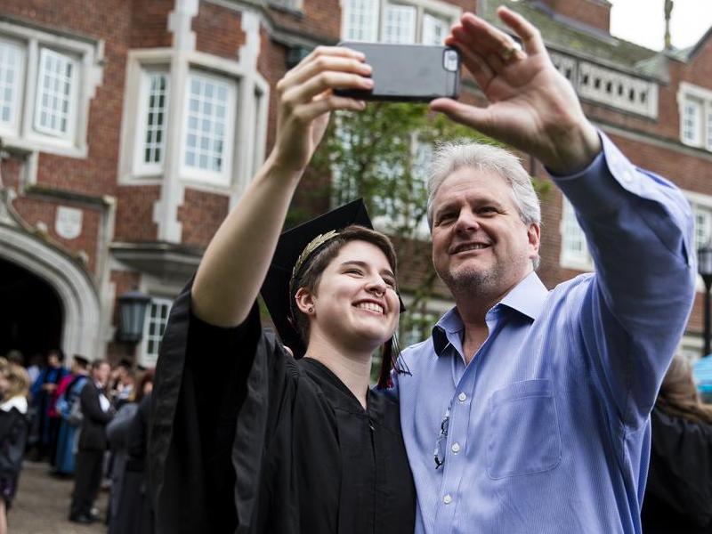 A student in graduation cap and gown and a parent take a selfie in front of Old Dorm Block.