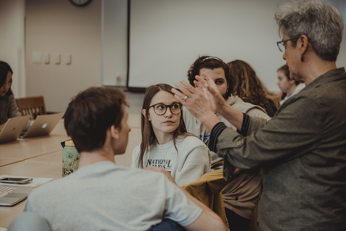 A professor stands behind a seated row of students and gestures with their hands while talking.