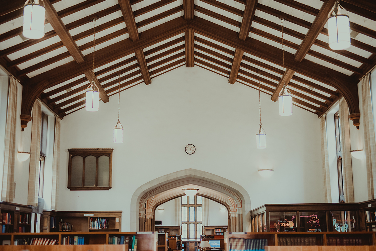A large room in a library with wooden bookshelves and cylindrical lamps hanging from a vaulted wooden beam ceiling.