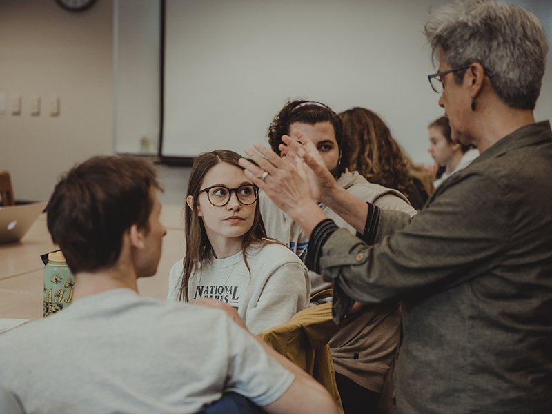 A professor stands behind a seated row of students and gestures with their hands while talking.