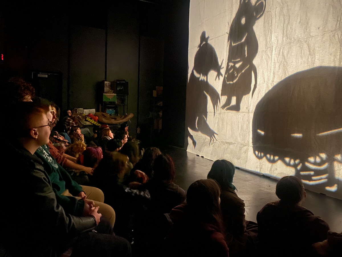 Audience watching a translucent skrim with images of mice in clothes projected as shadows on the skrim 
