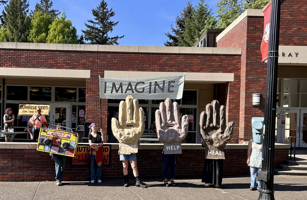 Performers wearing body-sized puppets of hands that read 'hope,' 'help,' and 'heal, along with a boombox that reads 'play' and a hammer, in front of a banner that reads "Imagine"