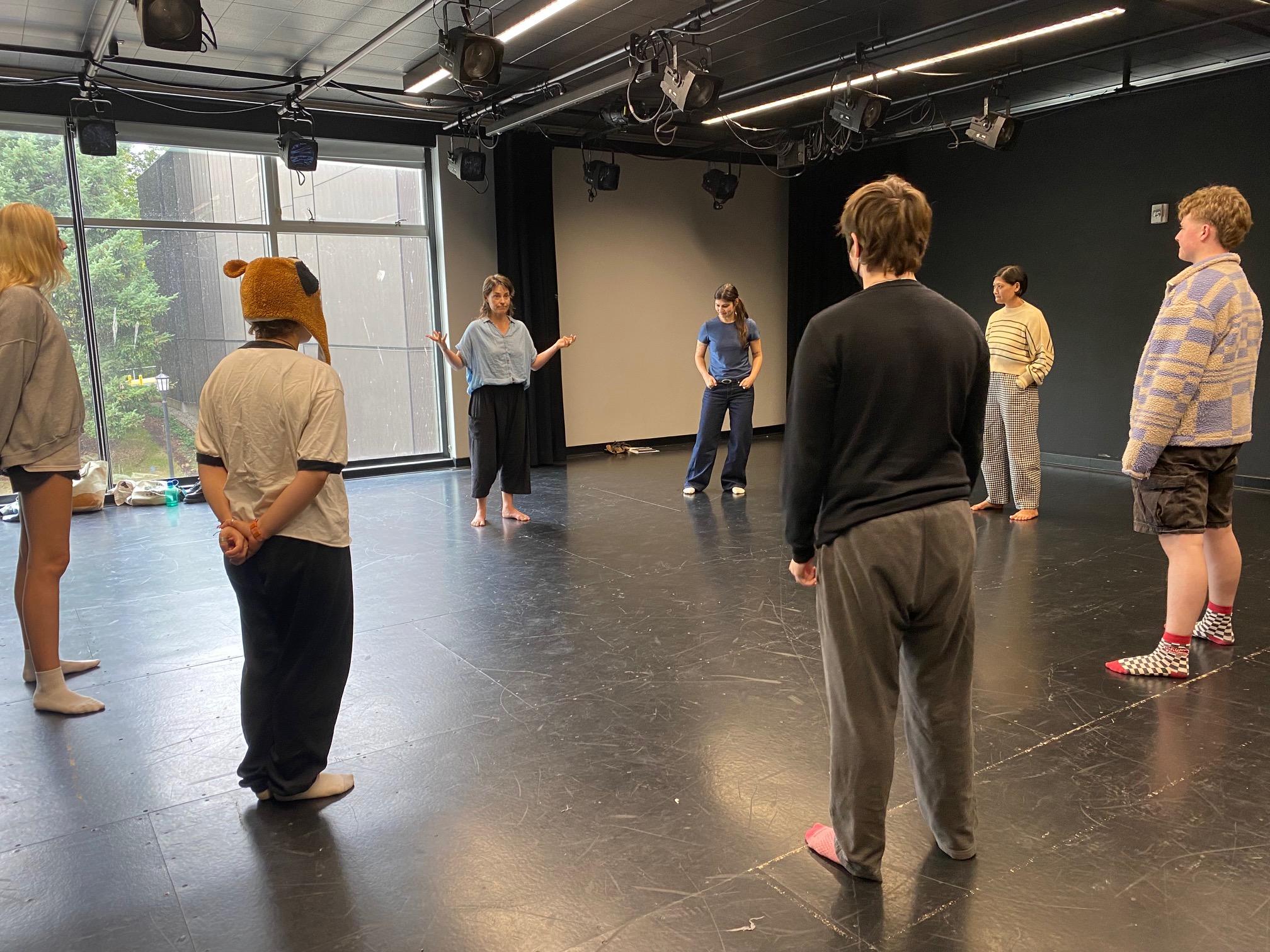 Two teachers stand in the middle of a circle of students in a blackbox room with plenty of space for movement.