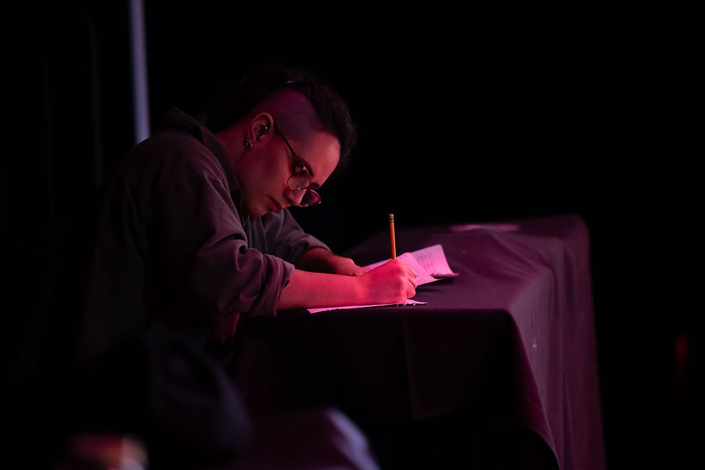 A student sits under a small light backstage writing notes on a piece of paper
