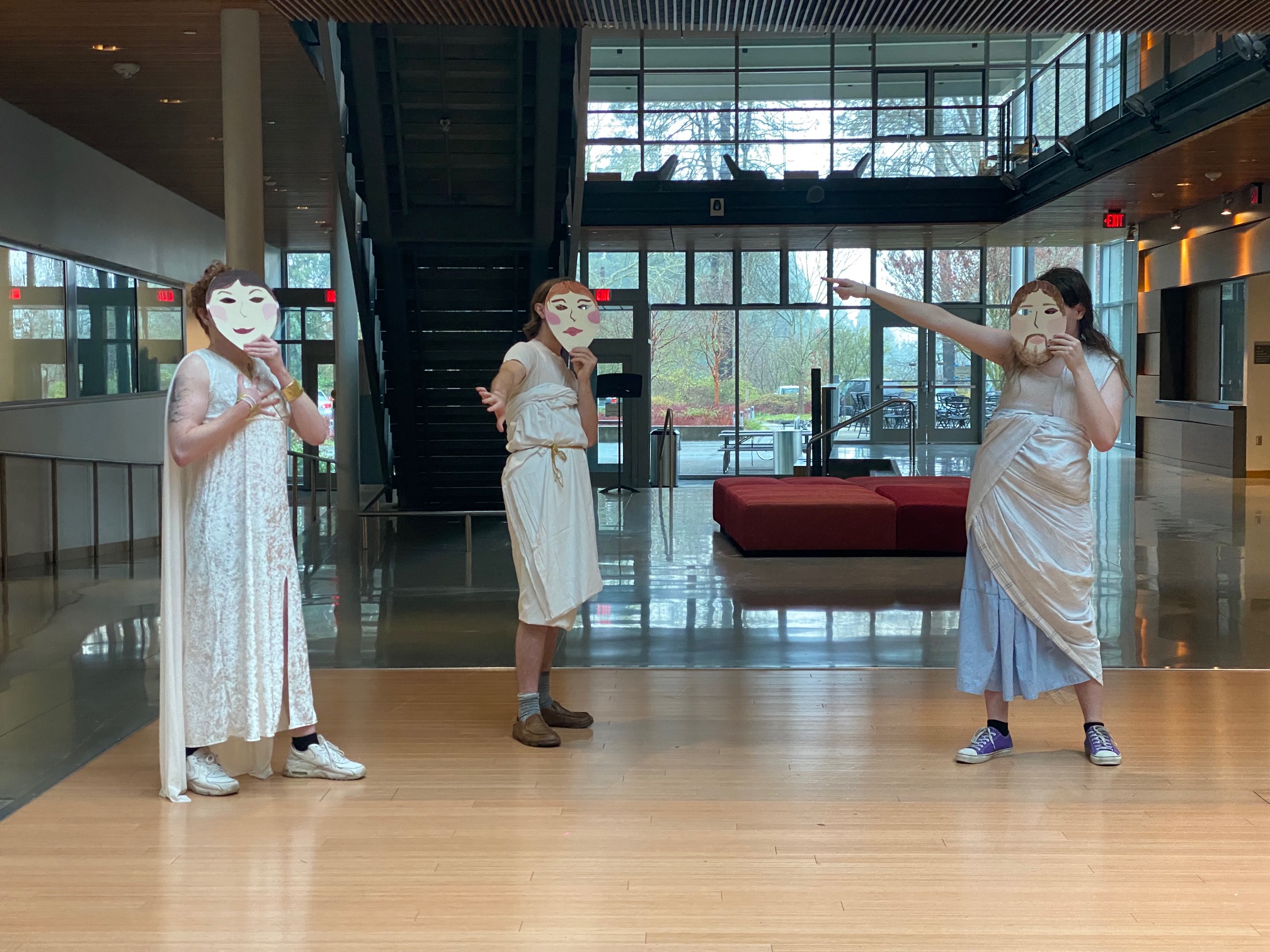 A group of three students pose dramatically in a well-lit indoor atrium in Greek togas and holding white masks over their faces.