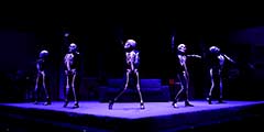 Five actors in skeleton costumes dance on a dark stage